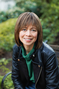 Headshot of Emily Midorikawa, a British-Japanese woman with short, dark brown hair and green eyes. She is wearing a green top and black leather jacket, and is sitting on a park bench.