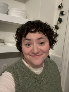 Sarah is a white woman with dark brown curly hair cut into a short bob with bangs. She is wearing a white t-shirt with a green hand knit vest