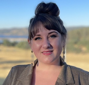 A brunette woman with her hair up in a bun and wearing a blazer smiles outside on a sunny day. The background is blurred. It’s a closeup of her face and shoulders.