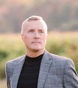 Headshot of Terrence P. Dwyer wearing a blazer and black turtleneck, with a soft-focus outdoor background.