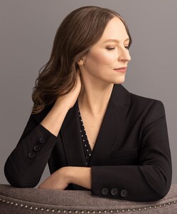 Photo of a White woman with long dark hair seated on an elegant, padded chair. She is facing the chairback, looking in profile to the right. She is only visible from the arms up. She wears an elegant black business suit.