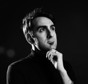 Black and white portrait of a man in a thoughtful pose, wearing a dark turtleneck, with hand resting on his chin against a dark background.