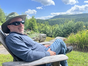 Man wearing green Tilly hat, blue sweatshirt, and sunglasses while sitting outdoors in an Adirondack chair with mountains in the background.