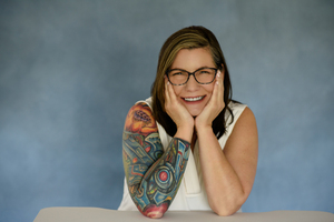 Portrait of Karen Marginot, wearing a sleeveless shirt, elbows on a table, with her hands on the sides of her face. She has a full-sleeve tattoo exposed.