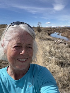 A woman with white hair and sunglasses on her head, wearing a blue shirt, in front of a creek.
