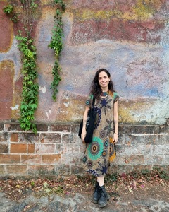 Robin Myers is standing against a stone wall with faded paint and hanging ivy. A thirty-something white cis woman, she has long brown hair and is wearing a woven necklace, a flower-patterned dress, and black boots. She smiles at the camera. 