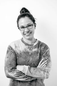 Black-and-white picture of a woman in a tie-dye, long-sleeve shirt, hair up in a messy bun.  She is smiling and wearing glasses, her arms are crossed.