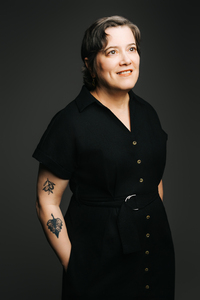 Portrait from waist up of woman with short brown hair, wearing belted black cloth dress with short sleeves