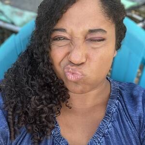 Headshot selfie of Samara Cole Doyon pursing her lips and scrunching her nose, playfully, twister her face a little to the right. Curly brown hair is draped over her right shoulder. She's wearing a denim sundress, with only shoulders and above visible.