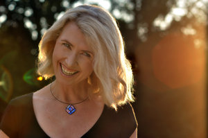 Backlit by sunlight, blond-haired, blue-eyed author Diana Dempsey is laughing in front of a backdrop of mature trees.