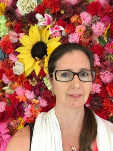 white woman with brown hair and wearing glasses and a white pajmina scarf in front of a wall of silk pink, red, and yellow flowers