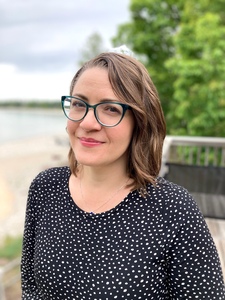 Profile photo of Alia Tawil: Smiling woman with shoulder-length brown hair and teal glasses, wearing a black dress with white polka dots, standing outdoors near a beach with trees in the background.
