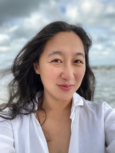 A middle-aged Asian American woman with dark silvering hair looks directly at the camera. She is wearing a white button down with a blurred background of blue and gray skies and seas.