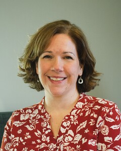Headshot of Rhonda, a white woman with chin length wavy brown hair, smiling wearing a patterned blouse against a light blue background.