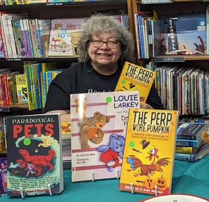 Smiling, frizzy-haired woman with glasses sits in front of bookshelves and holds book while other books are displayed on table.