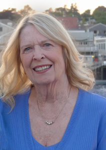 Older white woman with blonde hair to shoulders wearing a blue top in front of a small harbor 