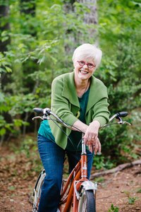 Photo of woman with white hair and a green sweater on an orange, old-fashioned bicycle, laughing.