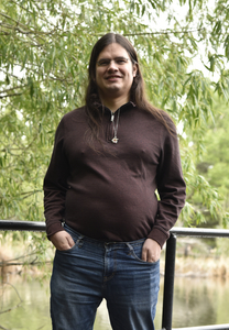 A man with some native American heritage wearing glasses, with long, dark hair standing next to a pond. He's wearing a maroon shirt and jeans.
