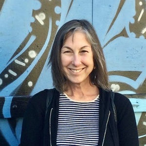 A woman in black sweater and striped shirt smiling against a blue wall mural.