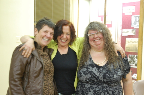 Three middle aged women are grinning in front of a lit library museum case. Hilary, in the middle in a lime jacket and black top, has her arms flung over the shoulders of the other two. 