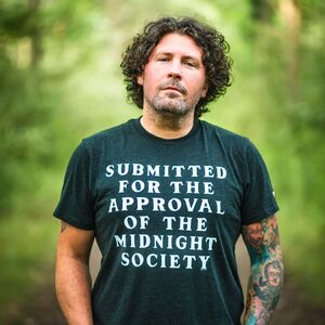 A photo of Michael Boulerice standing on a set of train tracks in the woods. Curly hair, black t-shirt that reads SUBMITTED FOR APPROVAL BY THE MIDNIGHT SOCIETY