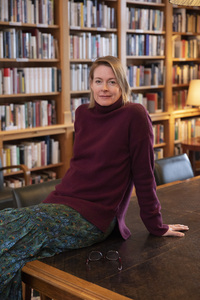 A 50 year old woman sitting on a table with a bookshelf behind her