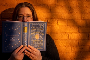 Image of me standing against a sun-lit orange brick wall holding up a copy of Alice's Adventures in Wonderland.