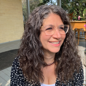 A white woman with brown curly hair in a black dress and glasses smiling outside