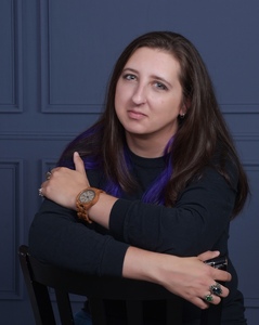 Half-body view of androgynous person sitting with arms crossed, long brown hair and blue sweater, blue background
