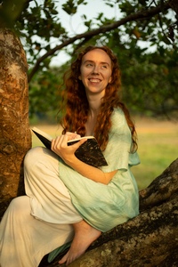 Anesce Dremen sits in a tree, smiling towards the sun while holding a notebook and pen 