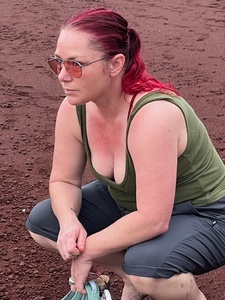 A middle-aged white woman squats on a red sand beach, gazing out into the distance. The photo is in profile. She has bright magenta hair caught up in a ponytail and wears rose-tinted aviators.