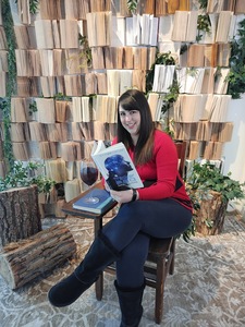My a white female in a red and black sweater with black leggings sitting at a desk reading my book TRACKER220 with a backdrop of books