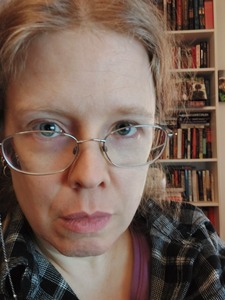 The author, a white woman with blonde hair & gray eyes (at least in this picture), stares intently at the camera through crooked bifocals. She is wearing a black and white plaid shirt over a bright lavender t-shirt. The bookcase behind her is overflowing.