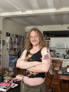 A white man with curly blond hair and a mustache smiles, crossing his arms, standing in a bright, high-ceilinged room with bookshelves and books on most surfaces. He wears gold-rimmed glasses and nose piercings. His arms have colorful tattoos.