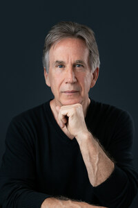 Author James L. Baker, wearing a black sweater with a neutral background, looking into at the camera with a quiet but intense expression