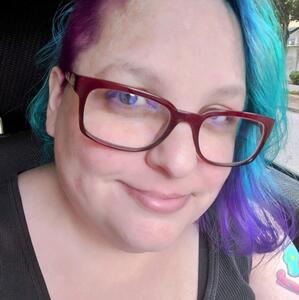 Photo of author & artist Raven Oak, sporting blue & purple hair and red-rimmed glasses.