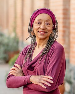 Author Jean Alicia Elster stands with her arms folded across her chest, next to a brick building, and with green shrubs in the background.