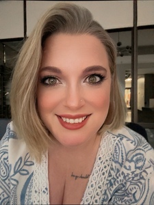 Author Dana Batista, a smiling white woman in her mid-forties with blonde shoulder-length hair, hazel green eyes, red lipstick, and a small nose stud, wearing a blue and white paisley top with white eyelet lace trim.
