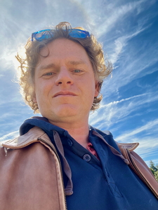 Photograph of author William Fertman, a middle-aged white man with curly hair, wearing an open leather jacket and blue hooded sweatshirt beneath. The photo was taken on a clear day with the sun directly behind him, creating a halo effect around his body. 
