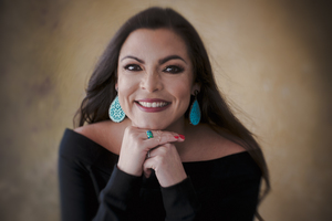 Portrait of Anna Orenstein-Cardona smiling, wearing turquoise earrings and a black top, with her chin resting on her hands.