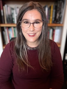 A photo of a woman with glasses, smiling. She sits in front of a bookshelf
