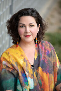 Image of Karla Helbert, Caucasian woman with dark hair, wearing bright colors and long beaded earrings in a casual pose looking up at the camera. 