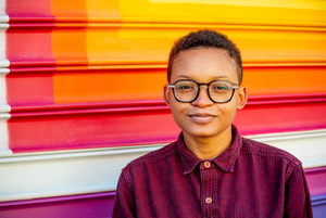 A black individual with angular glasses and short coily hair smiles. The background is colorful and they are wearing a burgundy button-up.