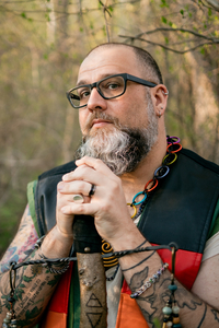 A middle-aged white man with arm tattoos. He has a bald head and a long beard that is gray, white, and brown. He has his hands crossed over a wooden staff. 