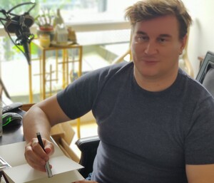 Alexander Paul Burton sitting at his desk in Toronto, Canada with a pen in his hand whilst smiling at the camera