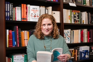 Author seated in a bookstore, holding a book and smiling softly, surrounded by shelves of poetry and nonfiction. She writes about the caregiver crisis and encourages caregivers to prioritize their own well-being.