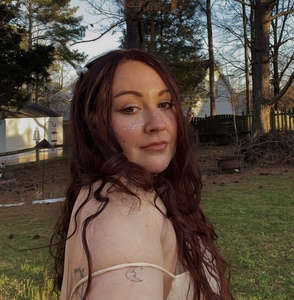 Photo of me, a woman with curly auburn hair, smiling at a camera with a wooded-area background and a cream colored dress on. 