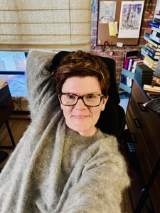 Portrait of an author with short auburn hair and tortoiseshell glasses, smiling while seated in a home office.