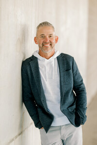 Author Brad Elder leaning casually against a light-colored concrete wall, wearing a charcoal blazer over a white hoodie, smiling warmly at the camera.