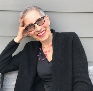 Author photo of Sarah Rosenthal wearing a black top, black sweater, pink beaded necklace, and glasses with black frames, sitting on a bench, leaning her head against her right hand, and smiling at the camera.